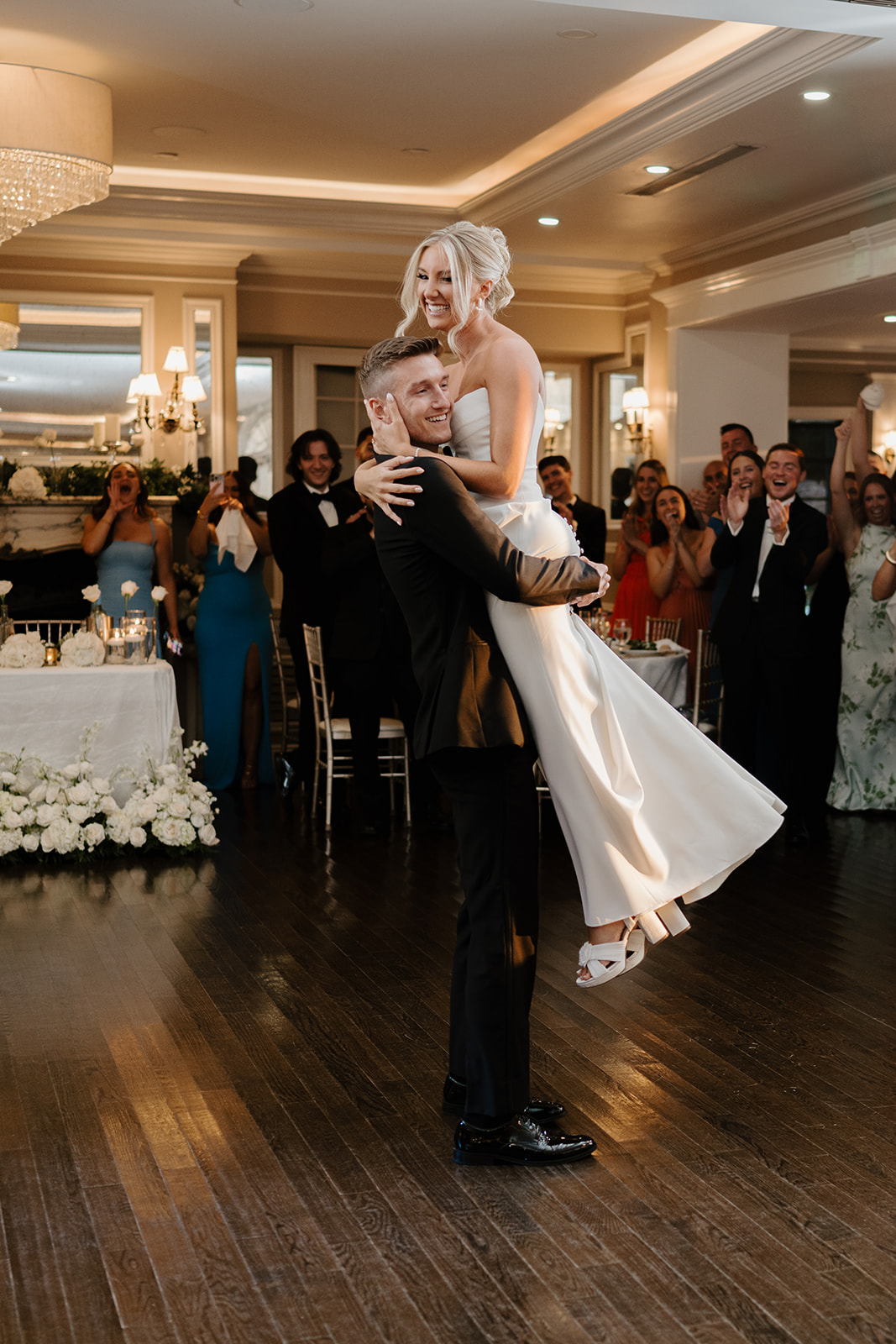 Groom lifts the bride during their first dance, surrounded by cheering guests in a warm-lit ballroom.